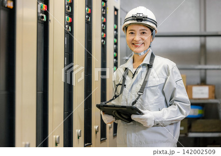 電子制御盤の前でタブレットを持つ作業服の女性 電子制御盤の前でタブレットを持つ作業服の女性 142002509