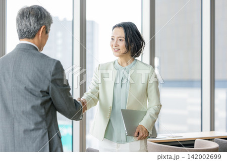窓際のオフィスで握手を交わすシニア男性とシニア女性 窓際のオフィスで握手を交わすシニア男性とシニア女性 142002699