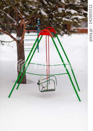 Green and red swing set stands in a snowy playground beneath a tree, surrounded by a blanket of fresh snow on the ground Green and red swing set stands in a snowy playground beneath a tree, surrounded by a blanket of fresh snow on the ground 142003312
