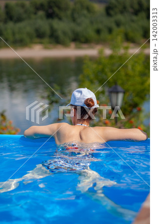 Young woman leans back in swimming pool, surrounded by blue water, with a river and greenery in the background, a lamp post nearby 142003313