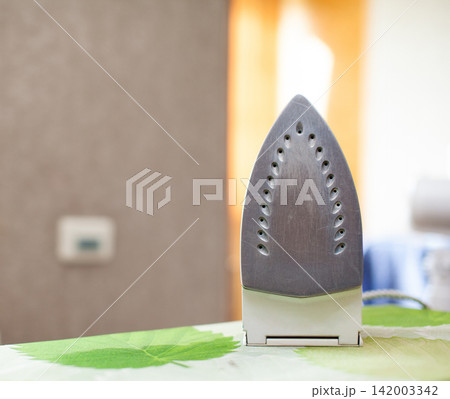 Iron positioned on a green patterned ironing board, with a softly blurred background showing a well-lit room and natural light filtering through a window Iron positioned on a green patterned ironing board, with a softly blurred background showing a well-lit room and natural light filtering through a window 142003342