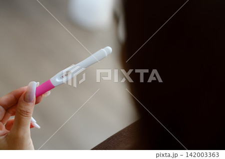 Woman's hand holds a pink and white pregnancy test, with a softly blurred indoor background featuring wooden flooring and neutral tones Woman's hand holds a pink and white pregnancy test, with a softly blurred indoor background featuring wooden flooring and neutral tones 142003363