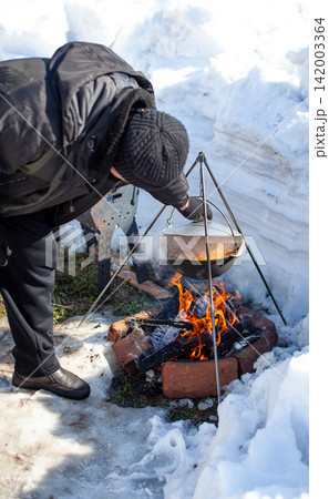 Individual prepares food over an open fire in a snowy environment, utilizing a tripod to suspend a pot above the flames Individual prepares food over an open fire in a snowy environment, utilizing a tripod to suspend a pot above the flames 142003364