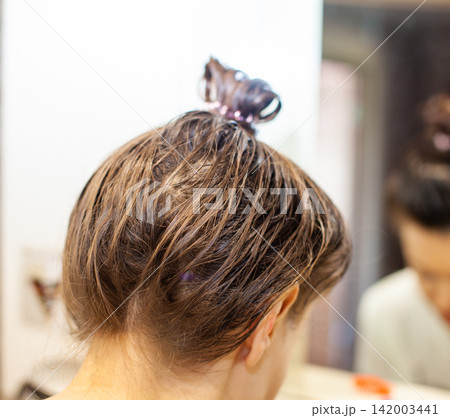 Woman with brown hair in a bun is seen from behind in a bathroom, with a mirror and soft lighting creating a serene atmosphere Woman with brown hair in a bun is seen from behind in a bathroom, with a mirror and soft lighting creating a serene atmosphere 142003441