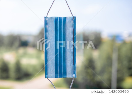 Blue surgical mask displayed outdoors, suspended by strings, with a blurred backdrop of greenery and distant structures under bright sunlight Blue surgical mask displayed outdoors, suspended by strings, with a blurred backdrop of greenery and distant structures under bright sunlight 142003490