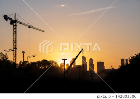 Silhouetted construction cranes against a colorful sunset, with a city skyline visible in the background, highlighting urban development and architecture Silhouetted construction cranes against a colorful sunset, with a city skyline visible in the background, highlighting urban development and architecture 142003506
