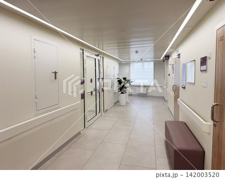 Modern hospital corridor showcases clean design with large windows, potted plant, and patient room doors, featuring a seating area along the walls 142003520