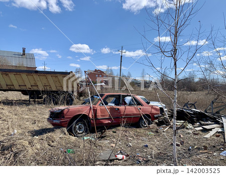 Abandoned red sedan rests in a field surrounded by debris and an old truck, with a clear blue sky and clouds overhead 142003525