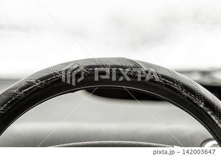 Close-up of a black leather steering wheel featuring a textured grip, with a blurred dashboard and vehicle interior in the background 142003647