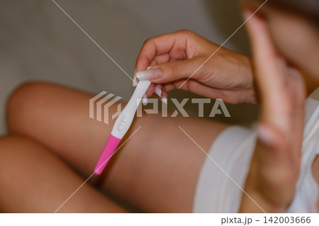 Woman sits on a bathroom floor holding a pregnancy test showing a positive result, dressed in a white dress, with a soft-focus background 142003666