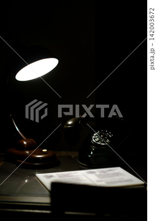 Vintage desk lamp casts light on a classic rotary telephone and an open book resting on a dark wooden table in a cozy environment 142003672