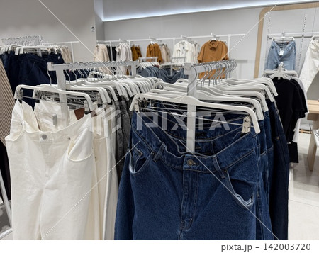 Assorted women's clothing items such as denim jeans and white pants are displayed on hangers in a contemporary retail store environment Assorted women's clothing items such as denim jeans and white pants are displayed on hangers in a contemporary retail store environment 142003720