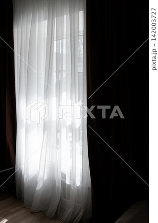 Sheer white curtains sway softly in sunlight beside dark brown drapes, creating a bright and airy atmosphere in an indoor space 142003727