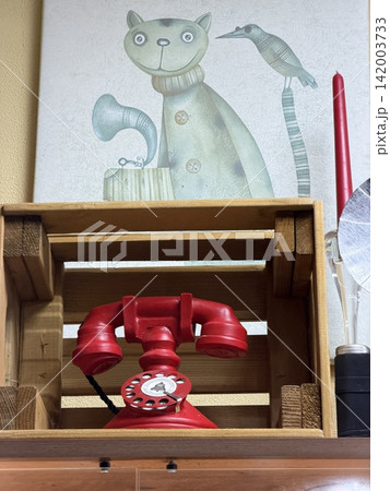 Kolomna, Russia - April 06, 2026: Vintage red rotary telephone sits on a wooden crate with a cat illustration and decorative items visible in the background 142003733