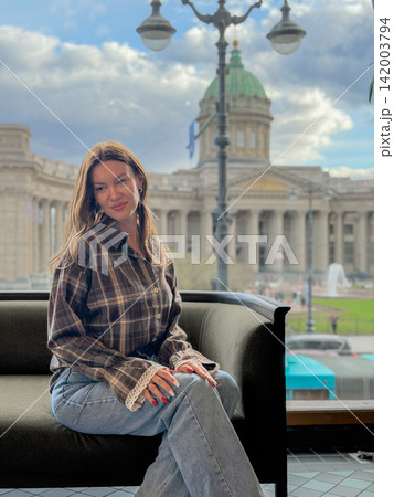 Woman wearing a plaid shirt and jeans sits on a sofa, with a historic building and cloudy sky visible through the window behind her Woman wearing a plaid shirt and jeans sits on a sofa, with a historic building and cloudy sky visible through the window behind her 142003794