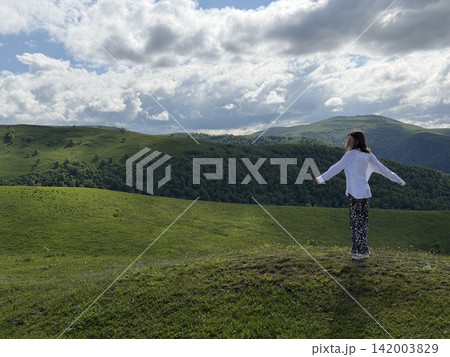 Young woman with arms outstretched stands on a hilltop, enjoying a panoramic view of lush green hills and a cloudy sky in the background Young woman with arms outstretched stands on a hilltop, enjoying a panoramic view of lush green hills and a cloudy sky in the background 142003829