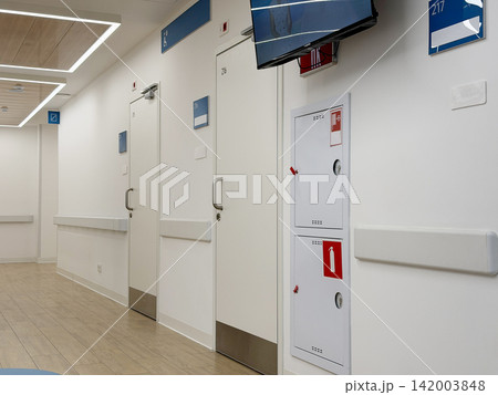 Hospital corridor displays numbered patient room doors and a wall-mounted television, showcasing a clean and modern healthcare environment Hospital corridor displays numbered patient room doors and a wall-mounted television, showcasing a clean and modern healthcare environment 142003848