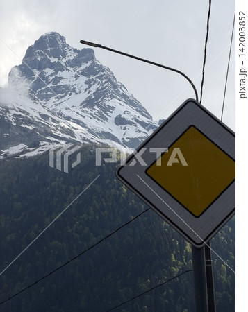 Snow-covered mountain peak towers over a lush green forest, with a caution traffic sign prominently displayed in the foreground Snow-covered mountain peak towers over a lush green forest, with a caution traffic sign prominently displayed in the foreground 142003852
