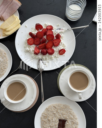 Breakfast table features cottage cheese with strawberries, oatmeal bowls, coffee cups, and a glass of milk on a dark surface Breakfast table features cottage cheese with strawberries, oatmeal bowls, coffee cups, and a glass of milk on a dark surface 142003915