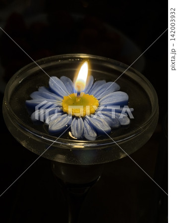Blue flower-shaped candle with yellow center is lit in a glass holder, casting a warm glow against a dark background with blurred objects Blue flower-shaped candle with yellow center is lit in a glass holder, casting a warm glow against a dark background with blurred objects 142003932