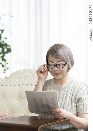 リビングで老眼鏡をかけて新聞を読むシニア女性 リビングで老眼鏡をかけて新聞を読むシニア女性 142010170