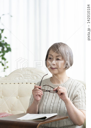 リビングで老眼鏡をかけて新聞を読むシニア女性 リビングで老眼鏡をかけて新聞を読むシニア女性 142010184