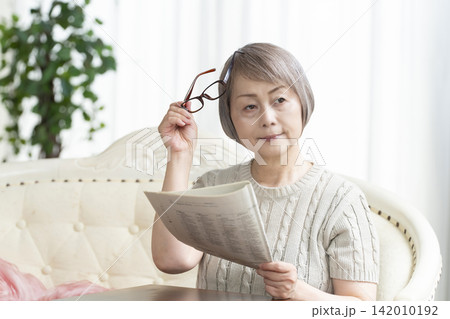 リビングで老眼鏡をかけて新聞を読むシニア女性 リビングで老眼鏡をかけて新聞を読むシニア女性 142010192