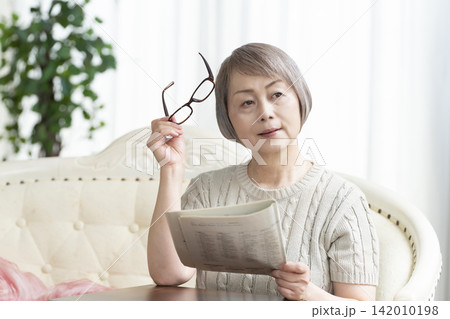 リビングで老眼鏡をかけて新聞を読むシニア女性 リビングで老眼鏡をかけて新聞を読むシニア女性 142010198