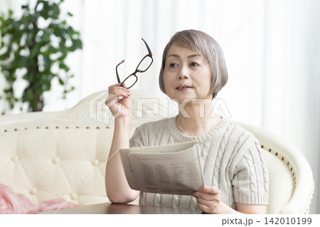 リビングで老眼鏡をかけて新聞を読むシニア女性 リビングで老眼鏡をかけて新聞を読むシニア女性 142010199