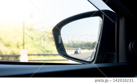Car side mirror capturing a clear view of an open road during daytime. Reflections in the rearview mirrors of cars driving along the asphalt road. With a long line of roadside barriers. 142010231