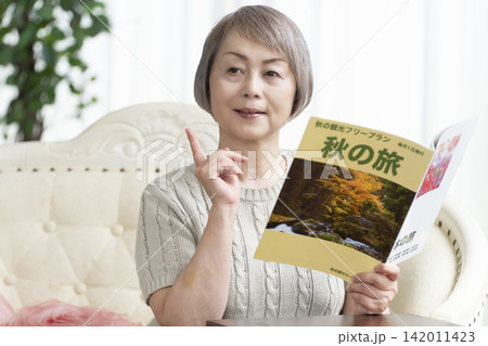 リビングのソファーで旅行雑誌を見るシニア女性 リビングのソファーで旅行雑誌を見るシニア女性 142011423