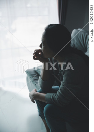 Thoughtful adult woman sitting alone by a bright window in a dark room, resting her hand near her mouth while worrying, overthinking, and facing an uncertain personal decision. Thoughtful adult woman sitting alone by a bright window in a dark room, resting her hand near her mouth while worrying, overthinking, and facing an uncertain personal decision. 142015488