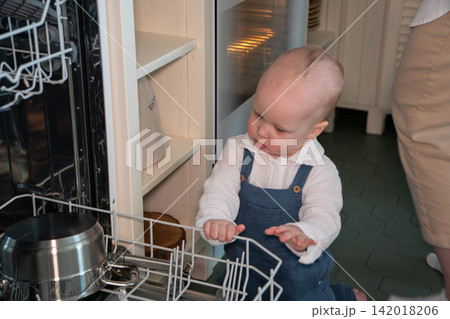 Baby stands beside open dishwasher and explores dish rack in home kitchen. Early childhood, curiosity, household safety, child development, exploration, learning, domestic life, everyday family Baby stands beside open dishwasher and explores dish rack in home kitchen. Early childhood, curiosity, household safety, child development, exploration, learning, domestic life, everyday family 142018206