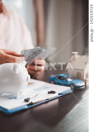 Piggy bank, blue car, wooden house, cash, and financial report show household budgeting, saving for major purchases, asset building, money management, and long-term financial goals. Piggy bank, blue car, wooden house, cash, and financial report show household budgeting, saving for major purchases, asset building, money management, and long-term financial goals. 142019724