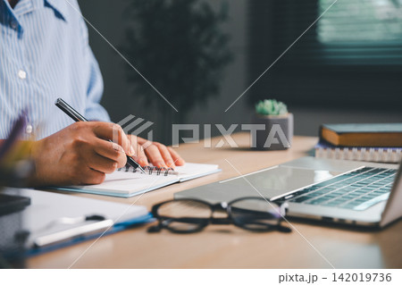 Person writing notes in a spiral notebook beside a laptop and eyeglasses at a home office desk, representing focused study, daily planning, remote work, and professional productivity. Person writing notes in a spiral notebook beside a laptop and eyeglasses at a home office desk, representing focused study, daily planning, remote work, and professional productivity. 142019736