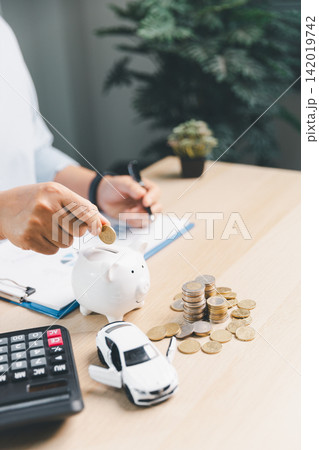 Person putting coin into piggy bank with calculator, white toy car, and coin stacks on desk, representing saving money for car purchase and auto loan planning. 142019742