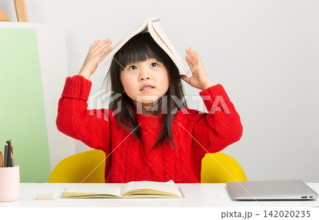 Playful Asian girl putting an open book on her head while sitting at desk. Playful Asian girl putting an open book on her head while sitting at desk. 142020235