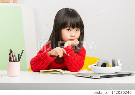 Bored little East Asian girl leaning on hand during home studying with laptop. 142020240