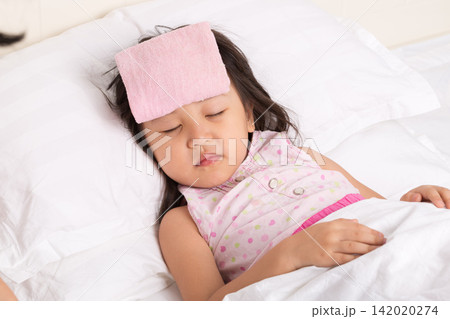 The sick little girl lay peacefully asleep in bed, a pink, damp towel on her forehead. The sick little girl lay peacefully asleep in bed, a pink, damp towel on her forehead. 142020274