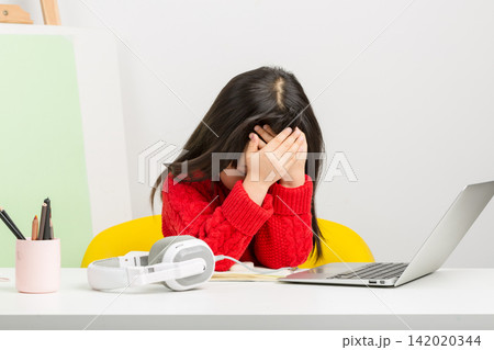 Frustrated East Asian schoolgirl covering her face with hands during online class at home. Frustrated East Asian schoolgirl covering her face with hands during online class at home. 142020344