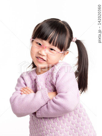 A cute little Asian girl wearing glasses stands smiling with her arms crossed in front of a pure white background. 142020360