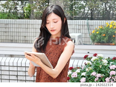 花壇のある屋外テラスでタブレット端末を持ち操作する若い日本人女性 142022314