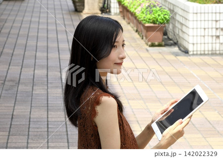 花壇のある屋外テラスでタブレット端末を持ち操作する若い日本人女性 142022329