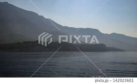 A serene coastal landscape viewed from a body of water, featuring a deep blue sea reflecting sunlight on ripples and peaks of gray mountains in a hazy misty atmosphere 142023601