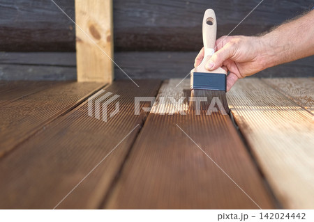 Brown oil for protection of wood by staining. Painting a larch for protecting and pests. Woodworker applying finish to terrace deck board. Repairman varnishing with brush wooden plank floor surface. Brown oil for protection of wood by staining. Painting a larch for protecting and pests. Woodworker applying finish to terrace deck board. Repairman varnishing with brush wooden plank floor surface. 142024442