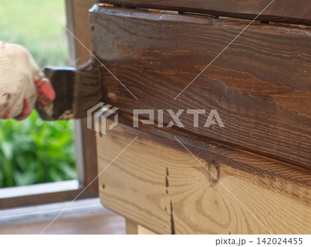 Brown oil for protection of wood by staining. Painting fence for protecting and pests. Woodworker applying finish to pine board box. Repairman varnishing with brush wooden fencing wall plank surface. 142024455
