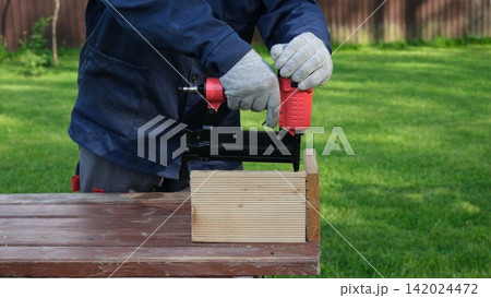 Cropped Image of Male Carpenter shooting nails on wooden frame outdoor. Industrial Cordless Nail Gun Power Tool. Tools efficient for framing, electrical. Carpenter using Air Nailer on timber. Woodwork 142024472