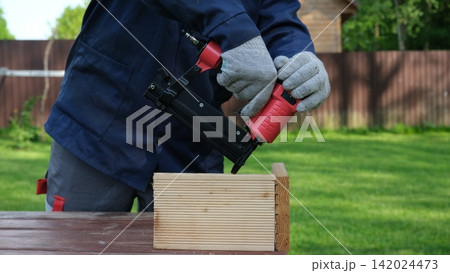 Cropped Image of Male Carpenter shooting nails on wooden frame outdoor. Industrial Cordless Nail Gun Power Tool. Tools efficient for framing, electrical. Carpenter using Air Nailer on timber. Woodwork 142024473