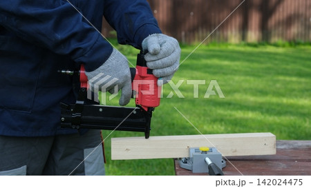 Cropped Image of Male Carpenter shooting nails on wooden frame outdoor. Industrial Cordless Nail Gun Power Tool. Tools efficient for framing, electrical. Carpenter using Air Nailer on timber. Woodwork 142024475