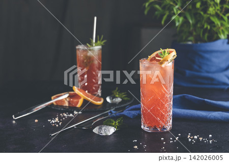 Pink Grapefruit Mezcal Paloma Cocktail in highball glass with water drops on table in real home. New life concept of quarantine, shallow depth of the field, dark background. Pink Grapefruit Mezcal Paloma Cocktail in highball glass with water drops on table in real home. New life concept of quarantine, shallow depth of the field, dark background. 142026005